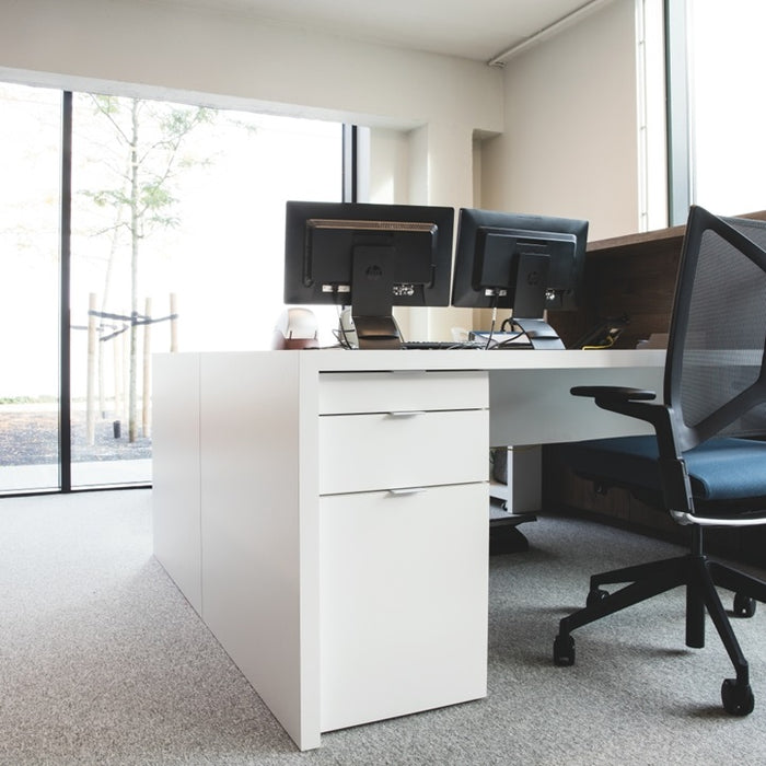 Modern office desk with computer monitors, a chair, and a filing cabinet in a bright room.